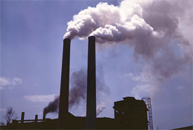 Two large smokestacks emit thick white and black smoke against a clear blue sky, with an industrial building partially visible at the base, illustrating the significant air pollution impacting the environment.
