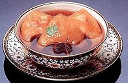 A decorative bowl filled with clear shark fin soup and three pieces of brown tofu, topped with a green garnish, is placed on a matching ornate saucer.