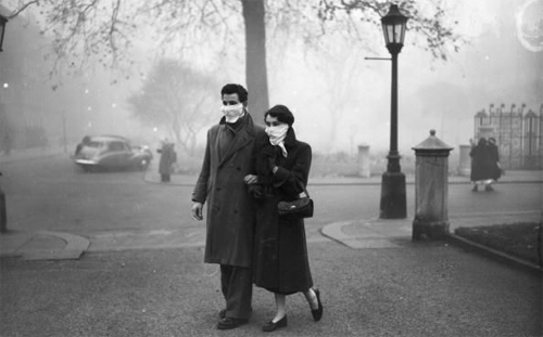 A couple strolls through a foggy park wearing masks, as vintage cars and pedestrians blur into the background, a stark reminder of the pervasive air pollution.