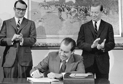 A man sits at a desk, pen poised, signing the Clean Air Act as two other men stand nearby, clapping. A painting hangs on the wall behind them.